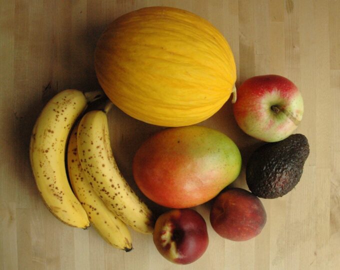 På et træbord ses lækre frugter i en bunke: Bananer, honningmelon, æbler, ferskner, avokado og mango.