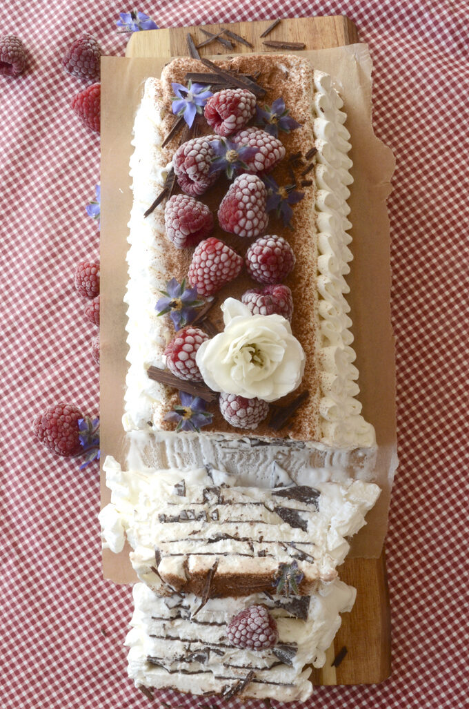 A vegan vienetta is seen from above, garnished with whipped cream swirls, frozen raspberries, white roses, chocolate thins and sugary cocoa powder. Two slices have been cut.