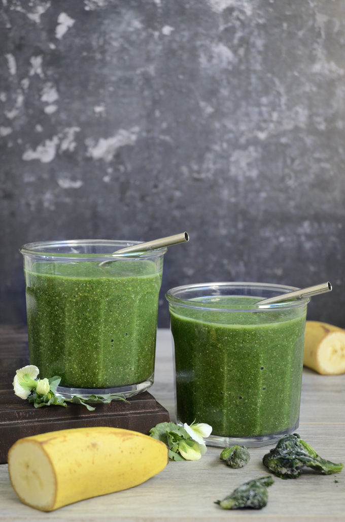 To glas med grøn smoothie. Der er sugerør af metal i hvert glas og rundt omkring ligger spinat og banan.