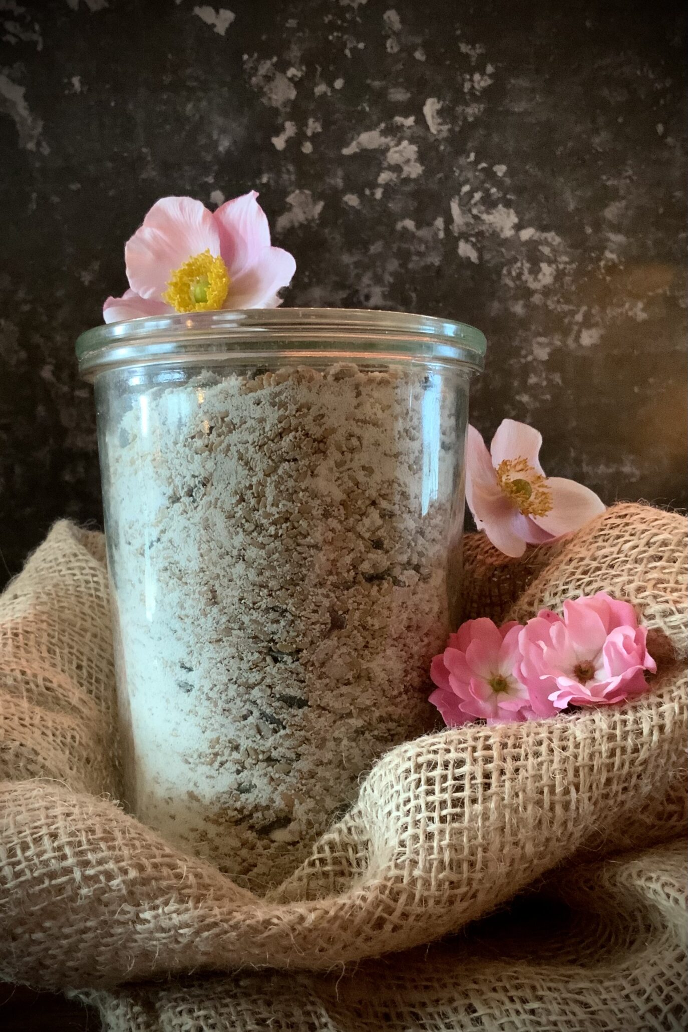 Et stort glas fyldt med nødder og kerner står på en hør-sæk. Omgivet af lyserøde blomster og med sort baggrund