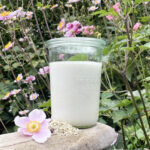 Omgivet af lyserøde blomster og grønne blade ses et stort glas med kridhvid hjemmelavet hampemælk stående på et stykke vejrbidt træ.